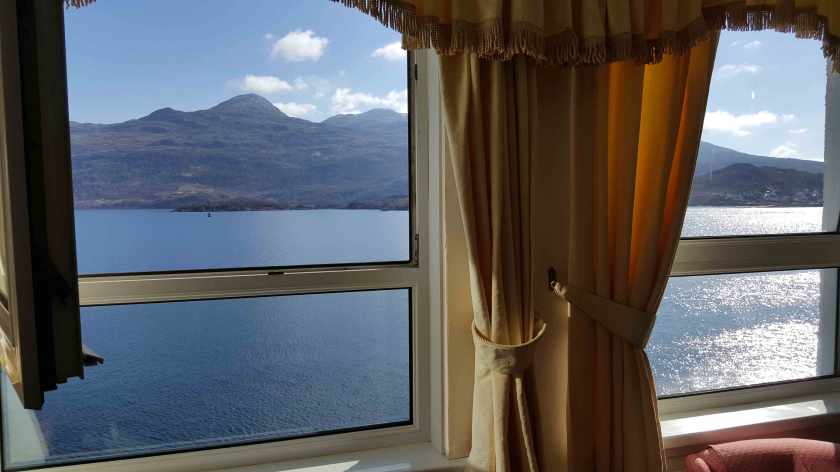 Past a curtained train window, a lake fronted by a mountain in the sunshine