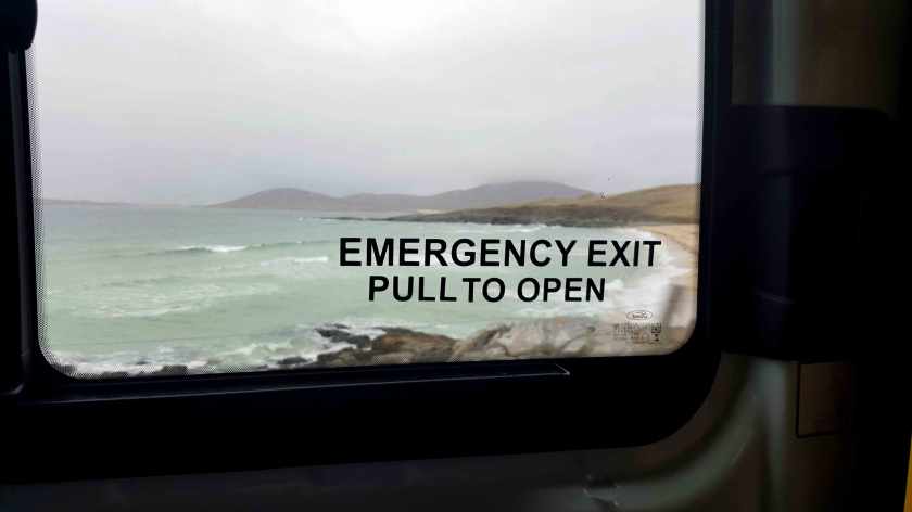 The view out the window of a vehicle of a beach. On the window the words Emergency Exit, Pull to Open.