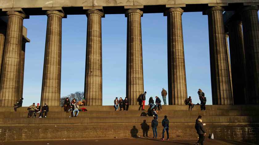 People sit on the portico of a neo-classical building against a blue sky.