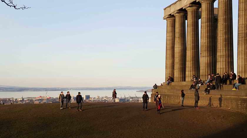 People sit in the sunshine beneath the portico of a Neo-Classical building at sunset. with the city in the distance.