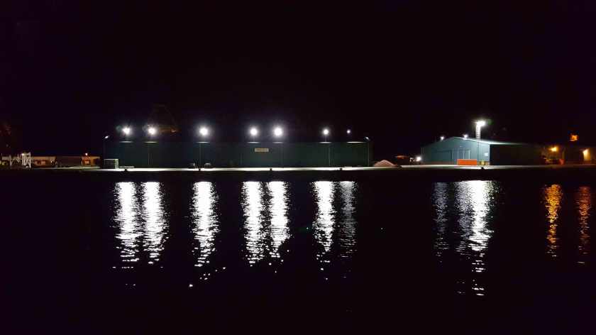 Lights from dockside buildings are reflected in the water at night.