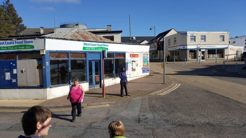A woman with white stick stands on the corner of a village street before the West Coast Food Store.