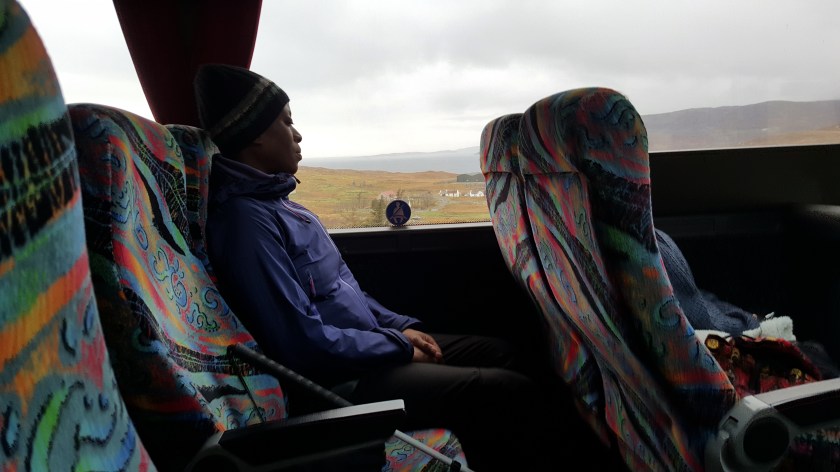 A woman travelling by coach leans her head against the window through which hills and a bay of water are visible.