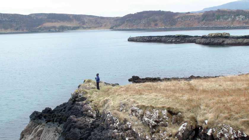 Seen from the distance and above, a woman holding a white stick stands at the edge of a cliff overlooking a bay