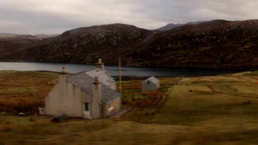 A small squarish house with slanted slate roof and an additional outbuilding beside a bay of water at the foot of a low mountain range