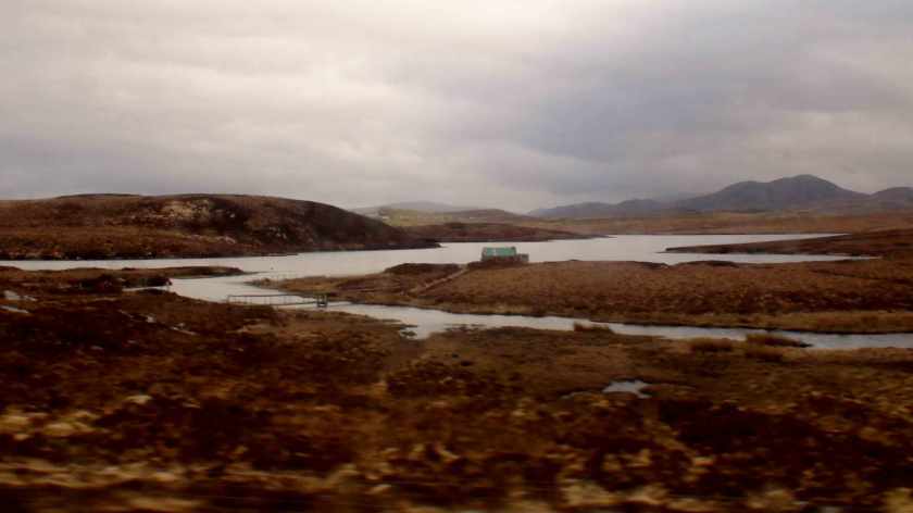 A tiny house in the distance sits at the edge of a bay of water in a dry orange landscape.