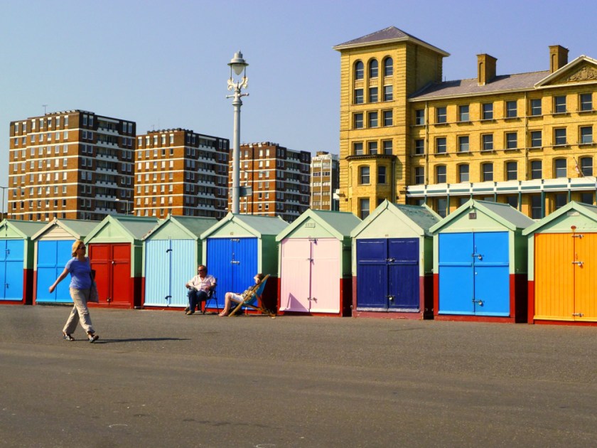 A woman walks past bright beach houses while a couple sits out on lawn chairs