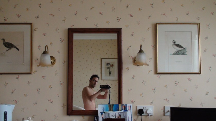 A bare-chested man takes a photo in the mirror of his hotel room