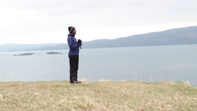 A woman stands, leaning on her white stick, at the edge of a bay.