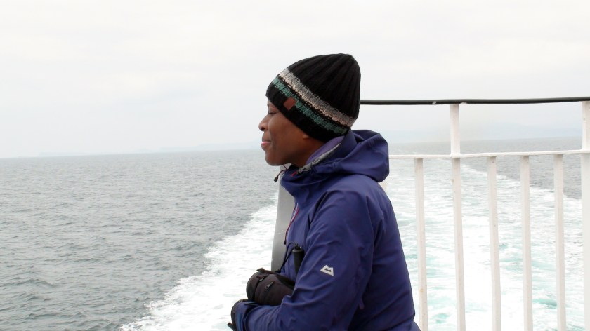 A woman leans against the rail of a ferry boat with the boat's wake visible behind her.