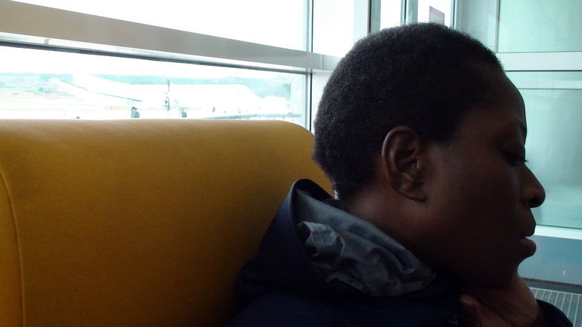 A woman sits on a mustard coloured bench with her head turned way, an airplane just visible out the window behind her.