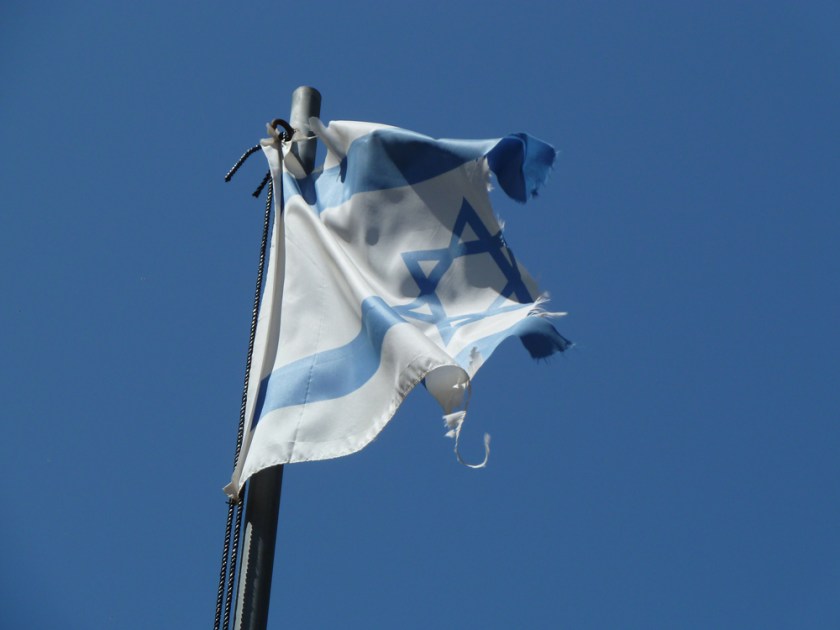 A tporn and ragged Israeli flag ripples against a blue sky