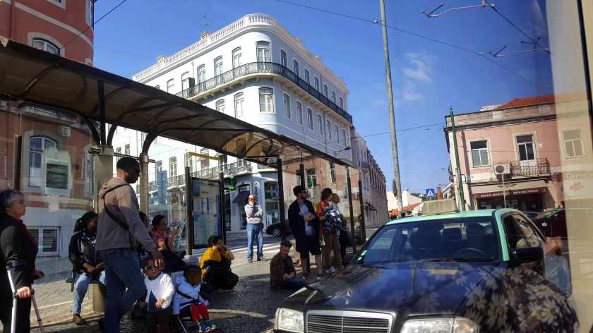 People gathered at a bus stop beside old European buildings and a passing car