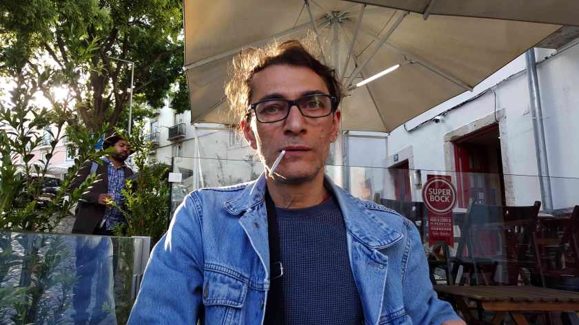 A man with glasses stares at the camera, a cigarette drooping from his mouth