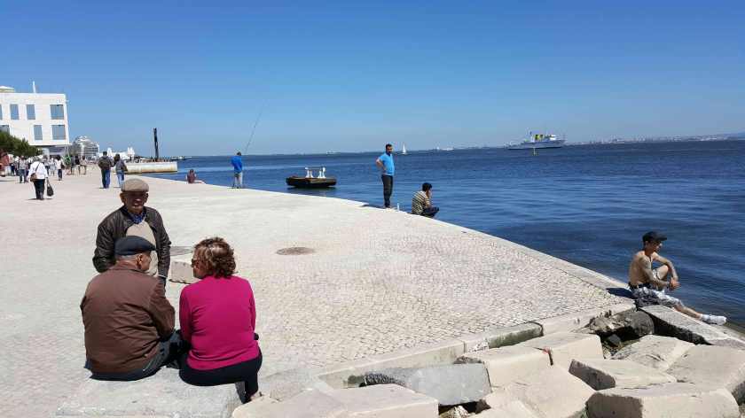 People sit at the paved edge of the water on a sunny day
