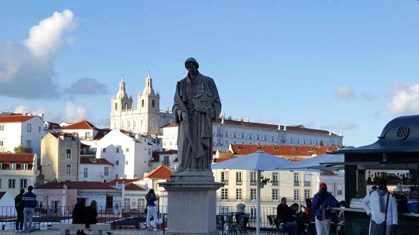 A stone figure stands on a pedestal in front of a hilly cityscape of old buildings including a large church on the hill