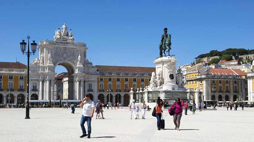 People walk through a city square with Baroque arch, building and statues