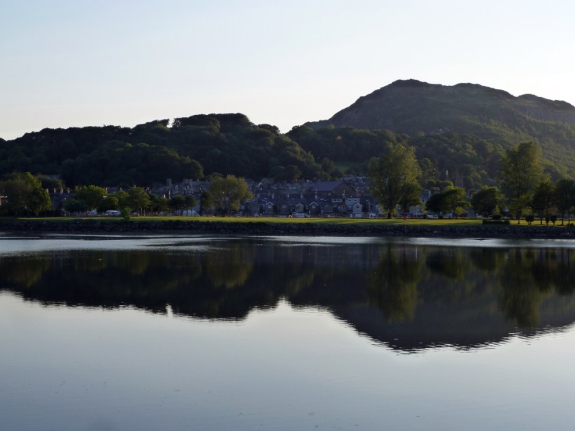 A village sits at the foot of a hill, both reflected in the glassy surface of the foreground lake