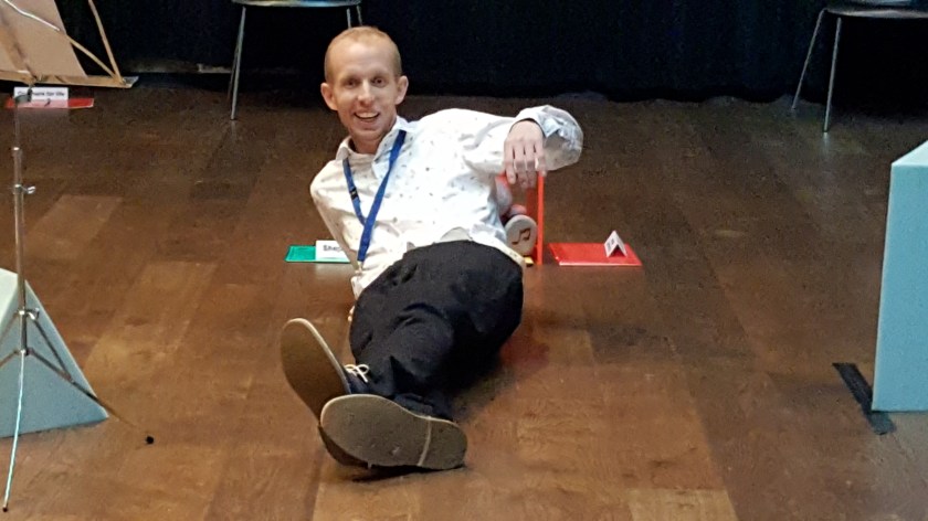 Tim lies on the floor with his left arm raised and grinning widely