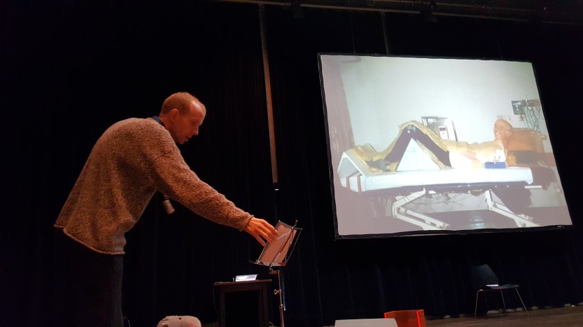 Tim adjusts pages on a music stand in front of a slide of himself with shaved head and raise legs on a hospital bed, grinning widely.