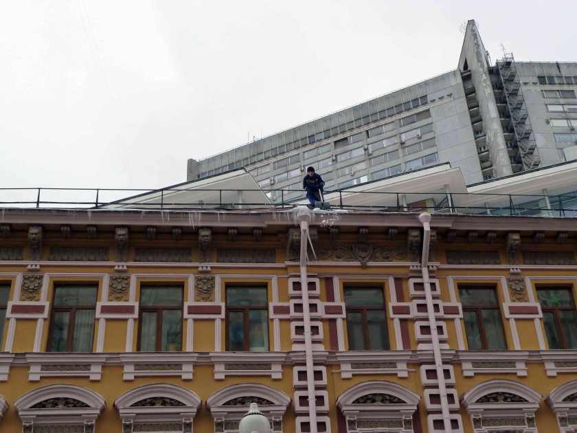 A man stands over the edge of a buidling's roof, shovelling snow
