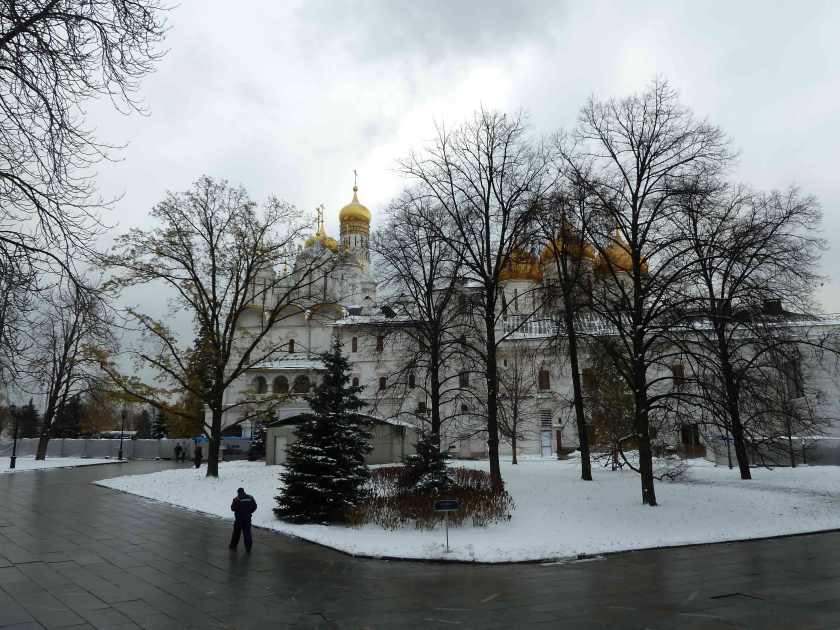 Bare trees stand in front of onion domed churches in a snowy landscape with a man in the foreground