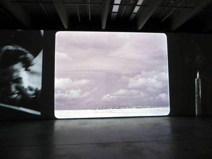 A screen installation in a museum shows a huge cloudy sky and, tiny in the distance, a long procession of people