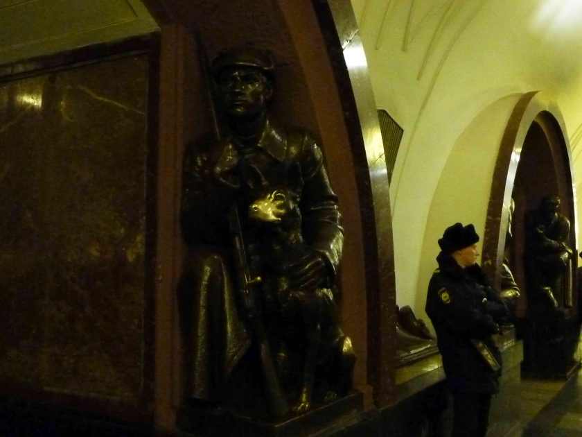 A soldier stands beside a USSR era bronze sculpture of a soldier kneeling beside dog.