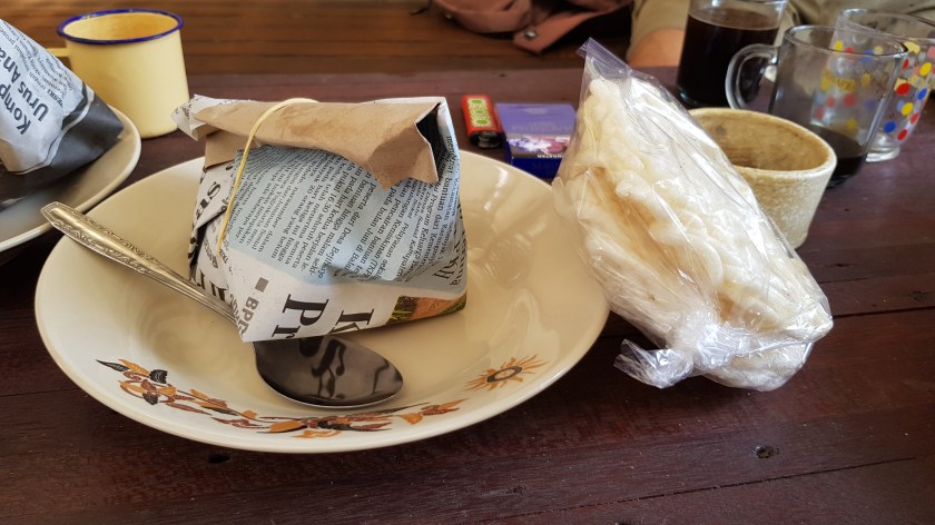 a folded newspaper package tied with string sits in a bowl beside a metal spoon, sided with plastic wrapped Indonesian rice cakes