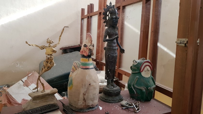 Four figures from different traditions on a table, a wire and plaster dancer, a painted hen, a bronze Indian classical dancer, and a painted ceramic frog.