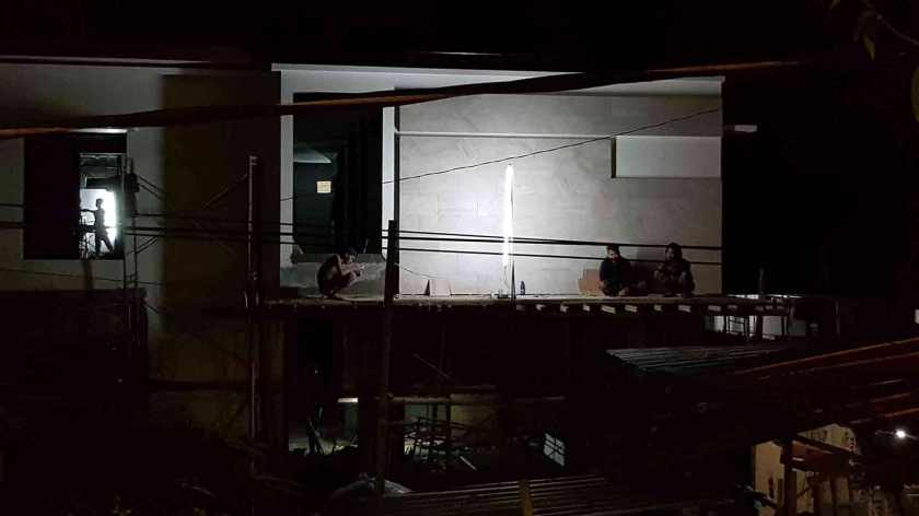 Lit by an electric lamp at night, three people sit on a deck outside a semi-constructed structure