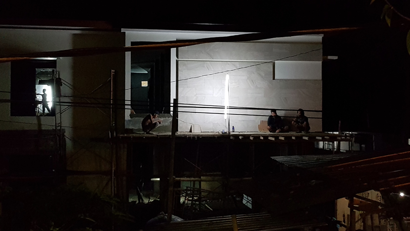Silhouetted by an electric lamp, figures rest on the balcony of a half constructed house. One squats, consulting his phone.