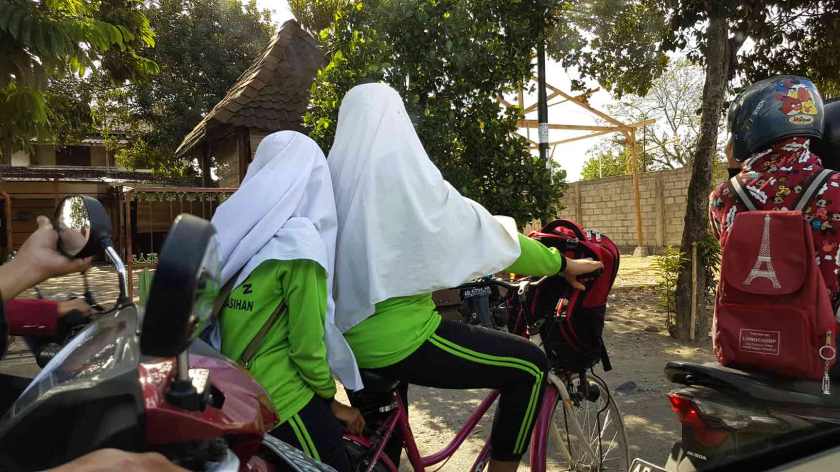 Two girls in hijab rest on a bicycle in the midst of Jogja traffic, only their backs visible