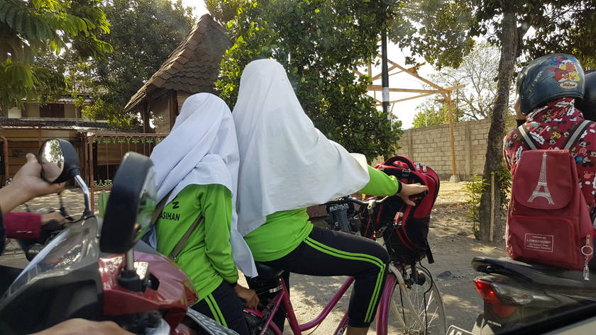 Two women in lime green gymsuits share a bicycle, their heads covered in white hijab and turned away, wedged between motorbikes.