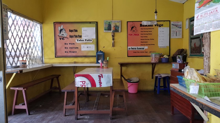 Interior of a warung, a small restaurant with picnic table and benches with the menu in Indonesian on the wall.