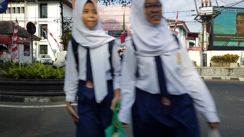 Blurred by their motion, two schoolgirls in uniform, including neckties and hijab, cross the road.