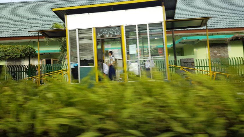 A youth stands at a bus stop, a small glass and metal booth, in school uniform, while bright blurred foliage rushes past in the foreground.