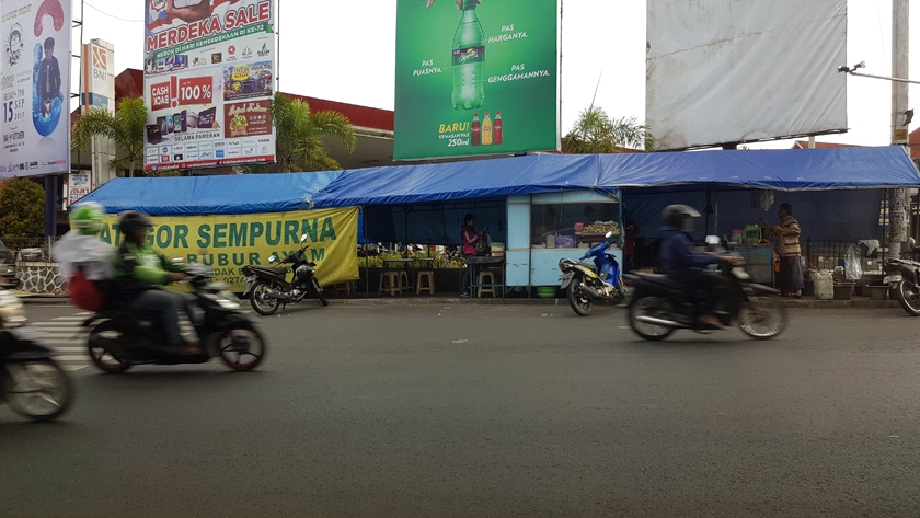 Urban street with food stall beneath giant billboards, at scooters pass in the foreground.