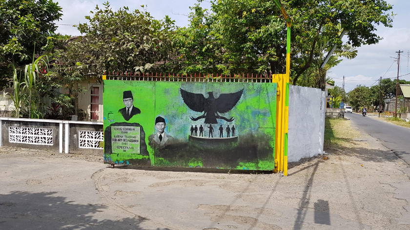 A mural painted on an open gate shows busts of Sukarno and Suharto beside the black silhouette of a spread-winged eagle hovering over a platform of small figures
