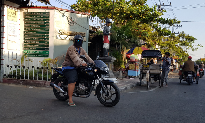 An androgynous motorcyclist wearing an opaque bubble helmet over their face rests in the road beside becak driver adjusting the awning of his vehicle