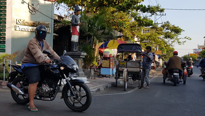 Large motorcyclist rests in the road with opaque bubble helmet beside a becak driver adjusting the awning of his vehicle