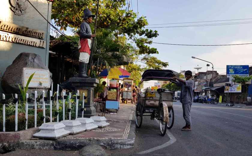 Smoking man fixes the awning of his three wheeled becak beside a lifelike wooden sculpture of a uniformed man holding a spear