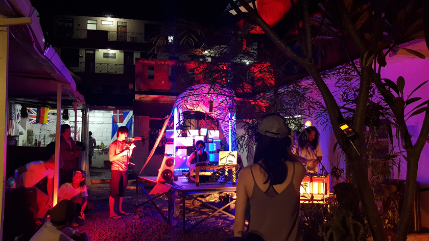 In the courtyard lit by purple and red light, band of electronic musicians and singer perform while one man films and others listen and watch