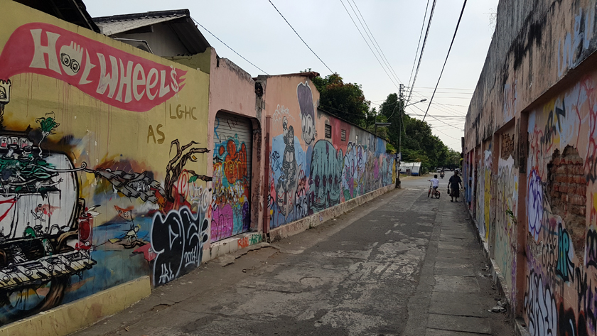 Jogjakarta backstreet, with colourful street art marking concrete walls and, in the distance, an older woman and a child on a bicycle.