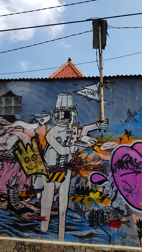 Jogjakarta street art: Surrounded by cartoon figures, a uniformed man with reflective face and steel hat carries a pole that extends into an actual street light.