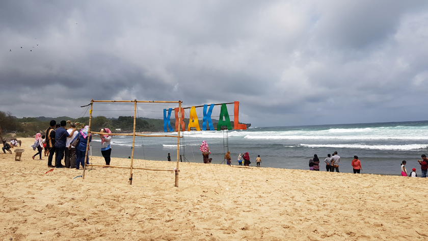Seashore with people at water's edge behind a handmade sign reading "Krakal"