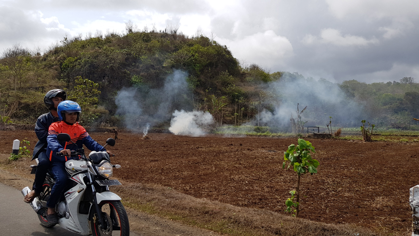 Two people on a motorbike pass a ploughed field with smoke billowing from burning waste