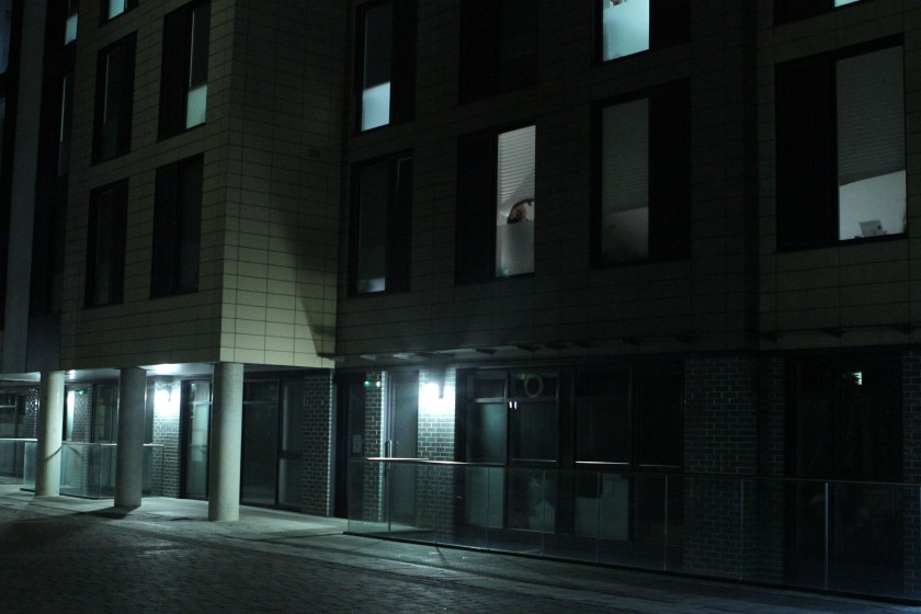 A figure peeks out through the blinds of a tower block at night.