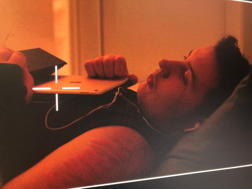 Orin lies in bed wearing headphones with ipad on their chest.