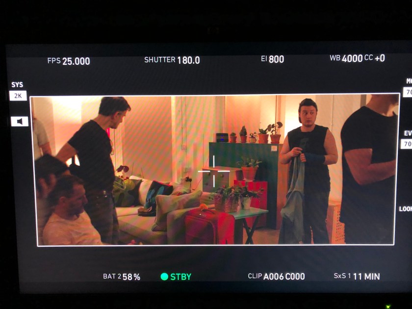 Actors and director on set in a living room with sofa and suitcase.